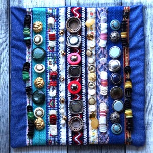 May include: A blue fabric panel with rows of colorful buttons, beads, and ribbons. The panel is decorated with a variety of textures and patterns, including stripes, polka dots, and floral designs.