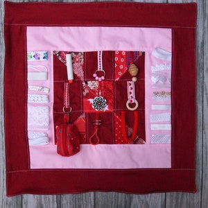 May include: A red and pink baby activity play mat with various textures and objects, including a zipper, a small pouch, a wooden ring, a rattle, and a variety of ribbons and laces.