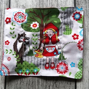 Könnte beinhalten: Quadratischer weißer Stoff mit einer farbenfrohen Illustration von Rotkäppchen, einem Wolf und einem Baum. Das Design umfasst rote, blaue und grüne Blumenmuster. Die Kanten sind mit rotem und blauem Stoff beschnitten.