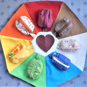 Puede incluir: Un círculo de tela colorido con un corazón en el centro y ocho pequeñas bolsas alrededor del borde. Las bolsas están hechas de diferentes telas y tienen varios patrones, incluyendo diseños florales, de lunares y geométricos.