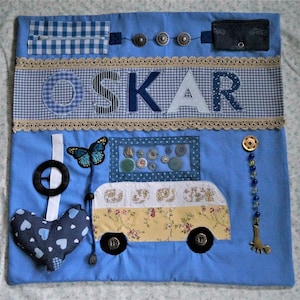 Puede incluir: Un tapete de actividades sensorial azul con varios elementos interactivos. Presenta un autobús con detalles florales, el nombre "OSKAR", botones, una mariposa, un corazón y un volante. Incluye una cremallera, adornos decorativos y un pequeño bolsillo.