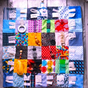 May include: A sensory activity board for babies and toddlers. The board is made of colorful fabric squares with various textures and interactive elements, including zippers, buttons, and ribbons. The board is designed to stimulate tactile exploration and fine motor skill development.