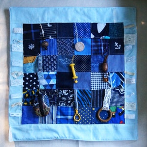 Puede incluir: Una manta sensorial azul y blanca con varias texturas y objetos, incluyendo un barco de madera, una cuenta de madera, un botón de madera, un juguete de plástico, un lazo de tela y un lazo de tela.