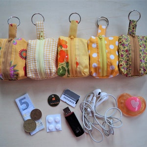 Könnte beinhalten: Fünf kleine, gelbe Stoffbeutel mit Reißverschlüssen und Schlüsselringen, jeweils mit einem einzigartigen Blumen- oder Musterdesign. Verschiedene Gegenstände wie Geld, ein USB-Stick und Ohrhörer sind ebenfalls abgebildet.