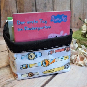 Puede incluir: Un contenedor de almacenamiento de tela blanca con una parte superior negra y un patrón de reloj colorido. El contenedor está lleno de libros, incluido uno titulado "Der erste Tag im Kindergarten" con un logotipo de Peppa Pig.