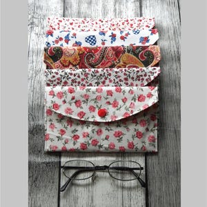 Könnte beinhalten: Fünf Stoff-Brillenetuis mit verschiedenen Blumenmustern. Das Etui vorne hat einen roten Knopfverschluss und einen weißen Hintergrund mit rosa Rosen. Die Etuis sind auf einer weißen Holzoberfläche gestapelt.