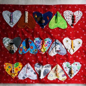 Puede incluir: Un cuadrado de tela roja con un estampado de lunares, con 12 parches de tela en forma de corazón. Cada corazón tiene un diseño único, incluyendo estampados florales, motivos de animales y varios adornos como cuentas y cintas. Los corazones están dispuestos en tres filas.