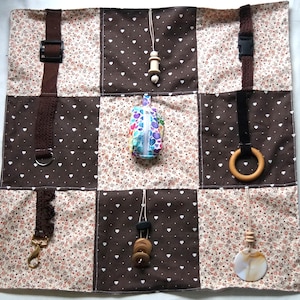 May include: A patchwork sensory mat with various textures and attachments. The mat features brown and floral patterned squares, with a variety of elements including a buckle, a D-ring, a clasp, wooden beads, a wooden ring, and a zippered pouch.