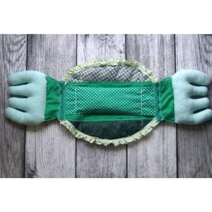May include: A handmade green and white polka dot baby pillow with two hand-shaped extensions. The pillow has a ruffled edge and is made of various green fabrics. The background is a wooden surface.