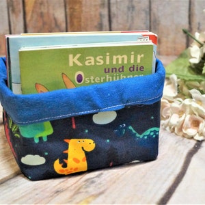 Può includere: Un cesto di stoffa blu con un motivo a stampa di dinosauro. Il cesto è pieno di una pila di libri per bambini. Il libro in cima ha il titolo "Kasimir und die Osterhühner".