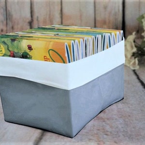 Puede incluir: Una caja de almacenamiento de tela gris con una banda blanca alrededor de la parte superior, llena de libros infantiles coloridos. Los libros tienen cubiertas amarillas, verdes y azules. La caja está sobre una superficie de madera.