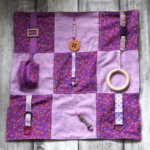 May include: A square fabric sensory mat featuring a grid of nine alternating purple polka dot and solid light purple panels. The mat includes various attached tactile elements such as a buckle, a small zippered pouch, wooden rings, beads, and ribbons.