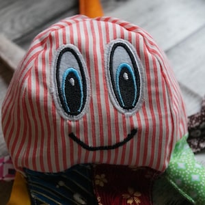 Puede incluir: Un juguete de peluche con una cara sonriente y una cabeza a rayas. El juguete tiene grandes ojos bordados con iris azules y apéndices de tela estampada. La cabeza es de rayas rojas y blancas.