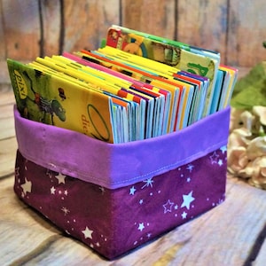 Könnte beinhalten: Ein lilafarbener Stoffaufbewahrungskorb mit weißen Sternen bedruckt, gefüllt mit einem Stapel bunter Kinderbücher.