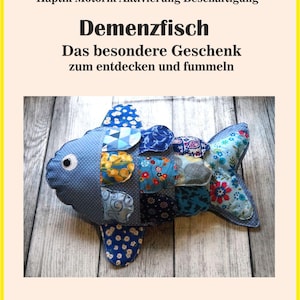 Puede incluir: Un juguete de pez de patchwork en tonos azules, con patrones que incluyen lunares y estampados florales. El juguete tiene un ojo blanco y está etiquetado "Demenzfisch" con texto alemán arriba y abajo. Sobre una superficie de madera.