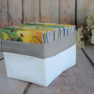 Könnte beinhalten: Eine weiße Stoffaufbewahrungsbox mit einem grauen Stoffbesatz, gefüllt mit einem Stapel bunter Kinderbücher. Die Bücher sind mit Illustrationen eines Frosches, eines Dinosauriers und anderer Tiere verziert.