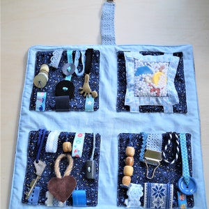 May include: A blue and white sensory activity quilt with four pockets. Each pocket is filled with different textures and objects, including ribbons, buttons, zippers, and beads. The quilt is designed to help children develop their fine motor skills and sensory exploration.