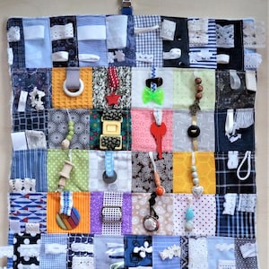 May include: A multi-colored fabric activity mat with various textures and objects for babies and toddlers to explore. The mat features different fabrics, ribbons, buttons, zippers, and other sensory items.