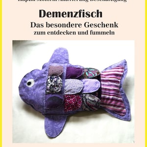 Może przedstawiać: Fioletowa zabawka w kształcie ryby z różnymi wzorzystymi łatami. Ryba ma duże oko i płetwy. Tekst na zdjęciu brzmi "Demenzfisch" i "Das besondere Geschenk zum entdecken und fummeln."