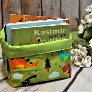 Può includere: Un cestino portaoggetti in tessuto verde lime con un motivo colorato di dinosauri, pini e nuvole. Il cestino è riempito con diversi libri per bambini, tra cui uno con il testo visibile Kasimir und die Osterhühner.