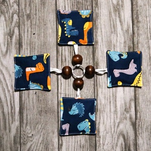 Puede incluir: Un juguete para bebés con cuatro cuadrados de tela azul marino con ilustraciones de dinosaurios en naranja, amarillo y azul. Los cuadrados están conectados por cuerdas blancas a una estructura central de cuentas de madera. Fondo de madera gris.