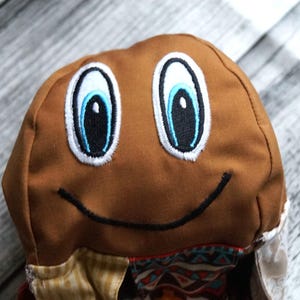 Puede incluir: Un sombrero de tela marrón con ojos de dibujos animados bordados y una boca sonriente. Los ojos son blancos con detalles azules y negros. El sombrero tiene un borde estampado.