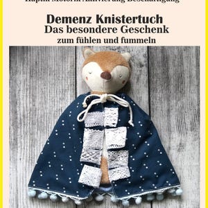 May include: A sensory blanket featuring a plush fox head attached to a dark blue fabric cape with white polka dots. The cape includes white lace squares, a white drawstring tie at the neck, and a light blue pom-pom trim along the bottom edge.