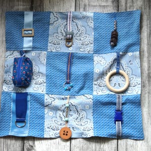 Könnte beinhalten: Eine quadratische sensorische Aktivitätsdecke mit einem Patchwork-Design aus hellblauem, strukturiertem Stoff und blauem Paisley-Stoff. Die Decke enthält verschiedene befestigte Gegenstände wie ein kleines Reißverschlusstäschchen, einen Holzring, eine blaue Schnalle und Perlen.