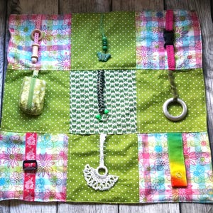 Puede incluir: Un tapete sensorial de patchwork con varias texturas y accesorios. El tapete presenta cuadrados de tela verde con lunares y flores. Incluye anillos de madera, una pequeña bolsa, un dije de mariposa y una hebilla. Diseñado para la exploración sensorial.
