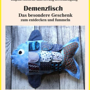 May include: A blue fabric fish toy with a variety of patterned patches, including paisley and geometric designs. The fish has a large eye and text that reads "Demenzfisch" and "Das besondere Geschenk zum entdecken und fummeln."