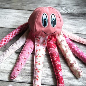 Puede incluir: Un juguete de peluche de pulpo rosa con ojos bordados y una boca sonriente. El pulpo tiene ocho tentáculos estampados en tonos de rosa y blanco. El juguete está hecho de tela suave.