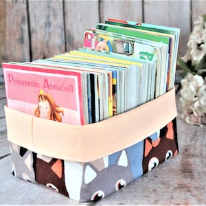 Puede incluir: Un contenedor de tela lleno de libros infantiles. El contenedor tiene un exterior estampado con caras de gato en gris, marrón y azul. Una banda color melocotón envuelve la parte superior. Las portadas de los libros son coloridas, con el texto "Prinzessin Annabell".