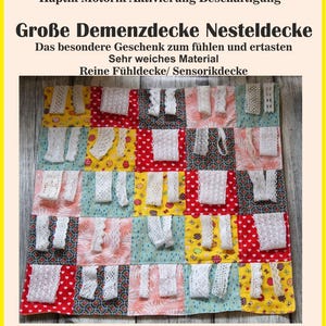 Könnte beinhalten: Eine quadratische Nesteldecke mit den Maßen 45 x 45 cm, bestehend aus bunten Stoffquadraten in Rot, Gelb und Blau. Jedes Quadrat ist mit weißen Spitzenbändern verziert. Der Text lautet: Demenz Autismus Alzheimer Haptik Motorik Aktivierung Beschäftigung Große Demenzdecke Nesteldecke Das besondere Geschenk zum fühlen und ertasten Sehr weiches Material Reine Fühldecke/ Sensorikdecke.