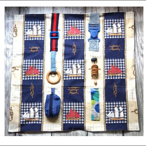 May include: A nautical-themed sensory activity mat with various textures and interactive elements. Features blue and white gingham squares with sailboat and knot designs. Includes a wooden ring, blue buckle, and a small blue pouch. The mat is designed for tactile exploration.