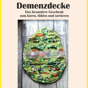 Op de afbeelding: Een groene sensorische deken met schildpad- en bladerpatronen, met diverse knopen, kralen en texturen. De deken heeft de tekst "Demenzdecke" en "Das besondere Geschenk zum hören, fühlen und sortieren".