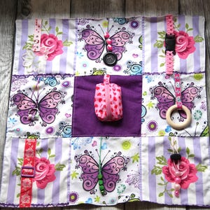 Puede incluir: Un cuadrado de actividades sensoriales para bebés y niños pequeños. El cuadrado presenta varias texturas, patrones y elementos interactivos. Incluye estampados de mariposas y flores, una pequeña bolsa con cremallera, botones, hebillas y un anillo de madera. La paleta de colores incluye morado, rosa y blanco.