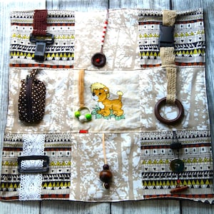 Könnte beinhalten: Ein mehrfarbiger Stoffquadrat mit verschiedenen Texturen und daran befestigten Objekten. Der Quadrat ist in neun Abschnitte unterteilt, jeder mit einem anderen Muster. Der mittlere Abschnitt zeigt einen Comic-Löwen. Weitere Objekte sind ein braunes Lederband, eine braune Reißverschlusstasche, ein Holzring, eine braune Holzperle, eine weiße Spitzenborte und ein brauner Knopf.