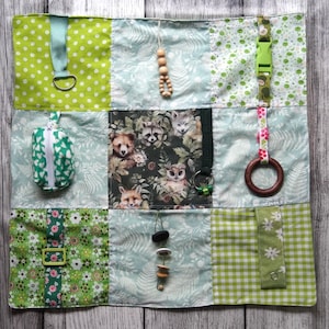 May include: A sensory activity mat with various textures and attachments. The mat features nine fabric squares in shades of green and blue, with polka dots, floral patterns, and woodland animal prints. Includes a zipper pouch, wooden beads, and a buckle.