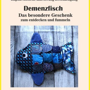 Puede incluir: Un juguete de tela con forma de pez azul y estampado, con el texto "Demenzfisch" y "Das besondere Geschenk zum entdecken und fummeln". El pez está hecho de varias telas azules y tiene un ojo blanco. La imagen también incluye las palabras "Demenz Autismus Alzheimer Haptik Motorik Aktivierung Beschäftigung" y "Beispielbild".
