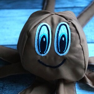 Puede incluir: Un peluche marrón con grandes ojos bordados en azul y blanco, y una sonrisa negra cosida. El juguete tiene largos apéndices marrones que se extienden desde su cabeza, lo que sugiere un diseño de criatura caprichosa.