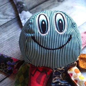 Puede incluir: Un juguete de peluche de pulpo hecho a mano con una cara sonriente. La cabeza es de tela a rayas verde azulado y blanca con ojos bordados y una sonrisa. Los tentáculos están hechos de varias telas estampadas, incluyendo diseños florales y geométricos. El juguete es para niños.