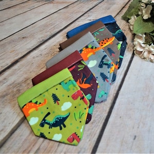 Könnte beinhalten: Ein Set aus sechs bunten Hundehalstüchern mit Dinosaurier-Druck. Die Halstücher sind aus Stoff und haben ein Farbschema in Grün, Braun, Grau, Rot, Blau und Dunkelblau. Die Halstücher zeigen verschiedene Dinosaurier, darunter einen T-Rex, einen Stegosaurus und einen Triceratops.