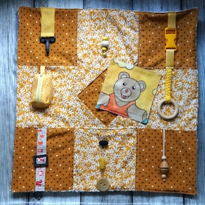 May include: A mustard-colored sensory board with various textures and interactive elements. Features include a small zippered pouch, a buckle, a wooden ring, a bear illustration, and a variety of beads and buttons. The board is designed for tactile exploration.