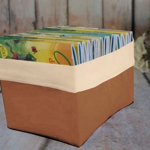 Könnte beinhalten: Eine rechteckige Aufbewahrungsbox mit zweifarbigem Design. Der untere Teil ist warmbraun, der obere Teil hat ein cremefarbenes Band. Die Box ist mit bunten Kinderbüchern gefüllt und bietet eine ansprechende Aufbewahrungslösung.