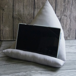 Könnte beinhalten: Ein grauer Stoffkissenständer für ein Tablet oder Buch. Der Ständer ist dreieckig geformt und hat eine flache Oberseite, auf der das Gerät liegen kann. Das Kissen ist aus einem weichen, bequemen Stoff.