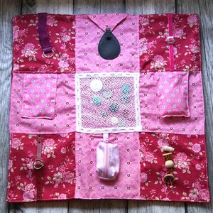 Puede incluir: Un tablero de actividades de tela rosa y roja con varias texturas y elementos interactivos. Presenta patrones florales, bolsillos, correas, botones y una pequeña bolsa. Diseñado para la exploración sensorial y el desarrollo de la motricidad fina.