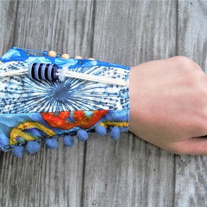 Puede incluir: Un brazalete de tela azul y blanco con un borde de pompón blanco y un acento de cuentas azul y blanco. El brazalete está en la muñeca de una persona.