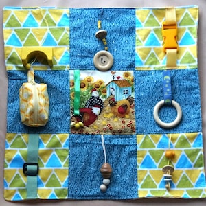 May include: A multi-colored baby activity play mat with various textures and features. The mat is made of fabric and has a variety of different patterns, including yellow triangles, blue triangles, and a blue and white pattern. The mat also has a variety of different textures, including soft fabric, smooth wood, and rough fabric. The mat has a variety of different features, including a zippered pouch, a wooden ring, a wooden bead necklace, a wooden button, a wooden toy, and a fabric loop.