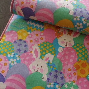 May include: A pink and green fabric table runner with a repeating pattern of Easter eggs and bunnies. The eggs are decorated with various patterns, including polka dots, stripes, and stars. The bunnies are white with pink noses and are peeking out from behind the eggs.