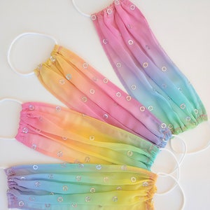May include: Four rainbow ombre face masks with white elastic ear loops and iridescent sequins. The masks are layered in a rainbow pattern, starting with pink, then yellow, green, and blue.